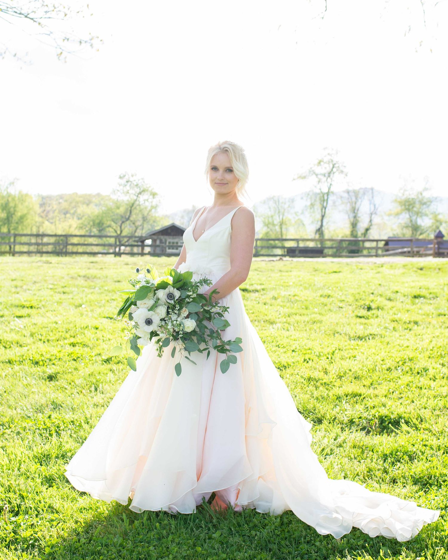 Bride in white V-neck wedding gown holding green and white bouquet standing on lush grass at outdoor farm venue.