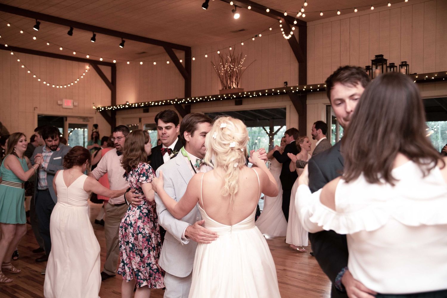 Bride and groom share a romantic dance surrounded by guests in a rustic barn wedding reception with string lights.