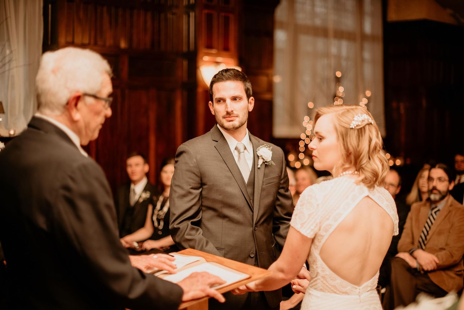 Bride and groom exchange vows at elegant indoor wedding ceremony with officiant and seated guests in background.