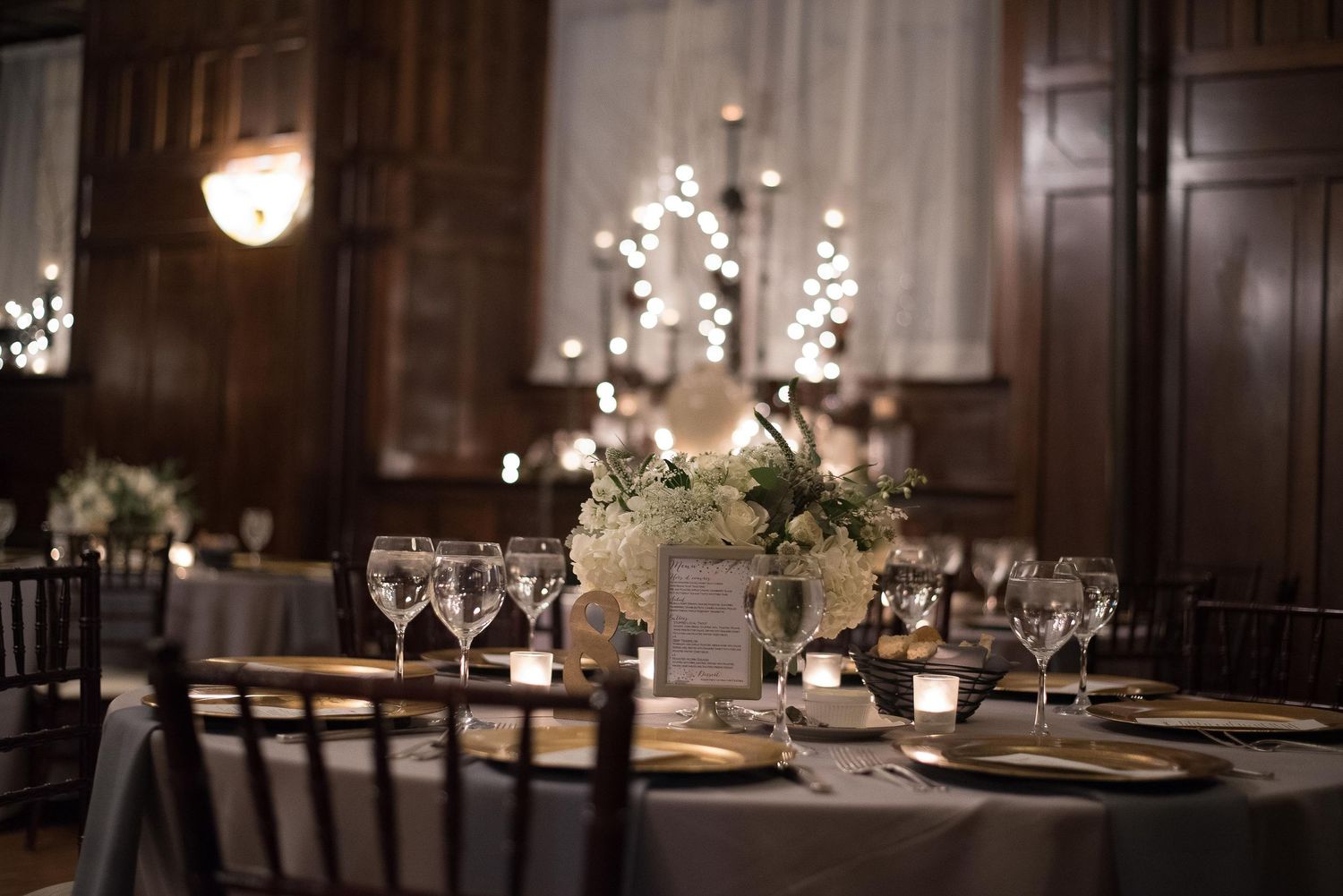 Elegant wedding reception table with white floral centerpiece, crystal glassware, and warm candlelight in a dark wood-paneled venue.