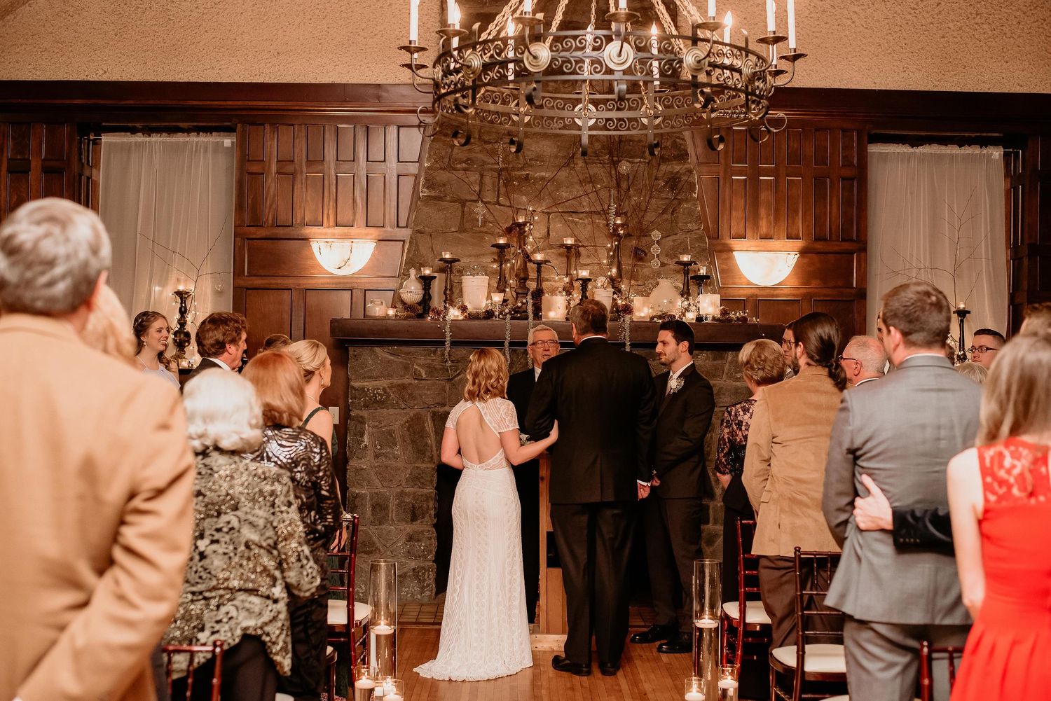 Elegant indoor wedding ceremony with bride and groom at stone fireplace altar, chandelier overhead, guests standing in rustic venue.
