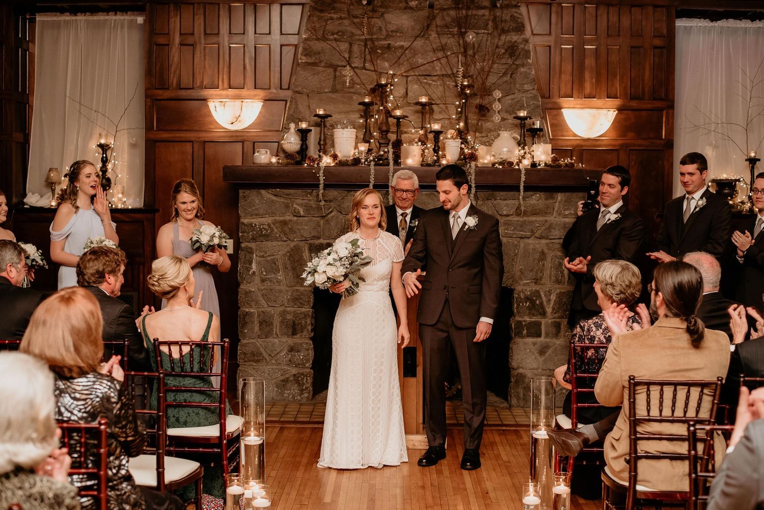 Bride and groom walk down the aisle at elegant indoor wedding ceremony with candlelight and stone fireplace backdrop.