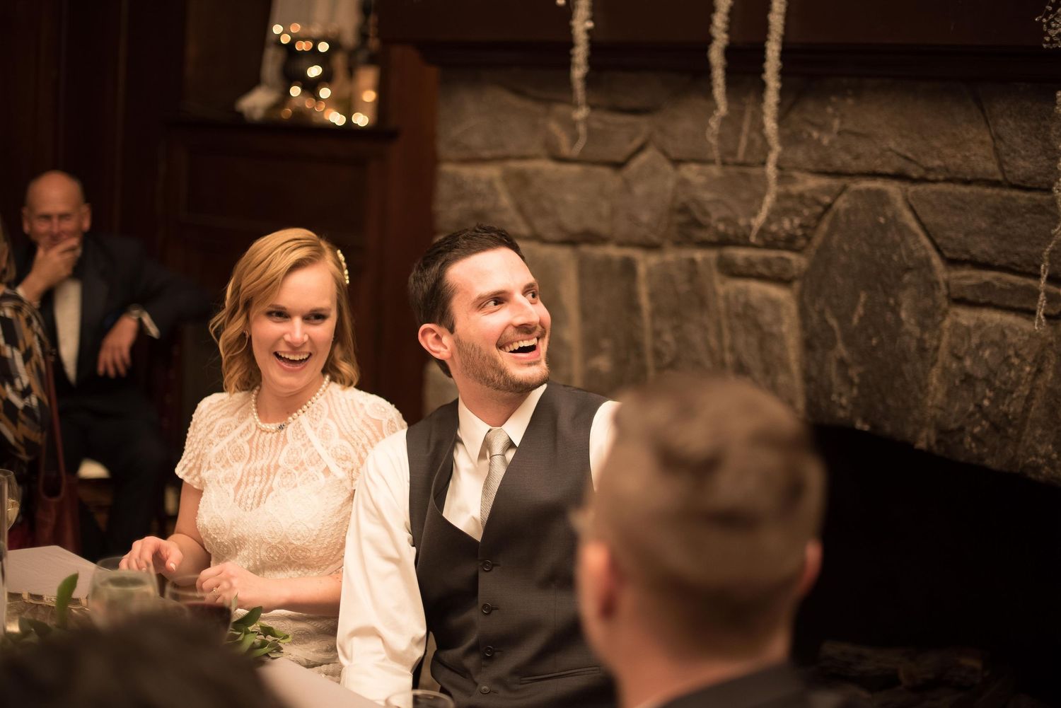 Happy couple smiling and laughing at their wedding reception seated in front of a rustic stone fireplace.