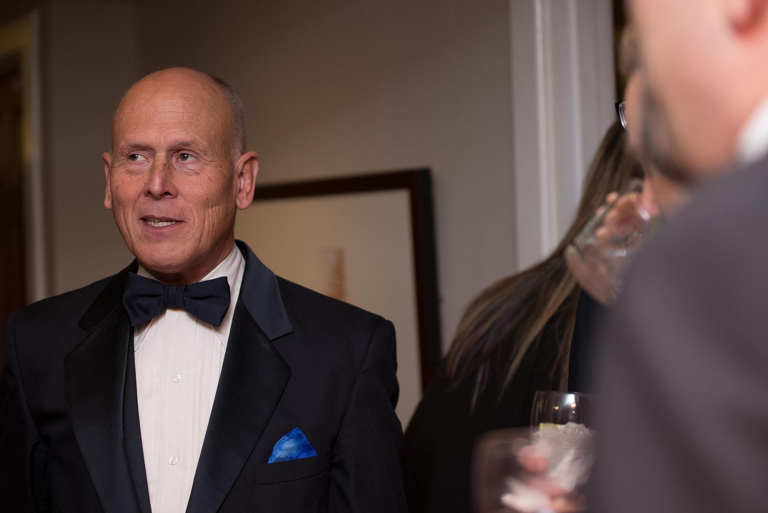 Elegant man in tuxedo with blue pocket square at formal event, socializing with guests in dimly lit venue.