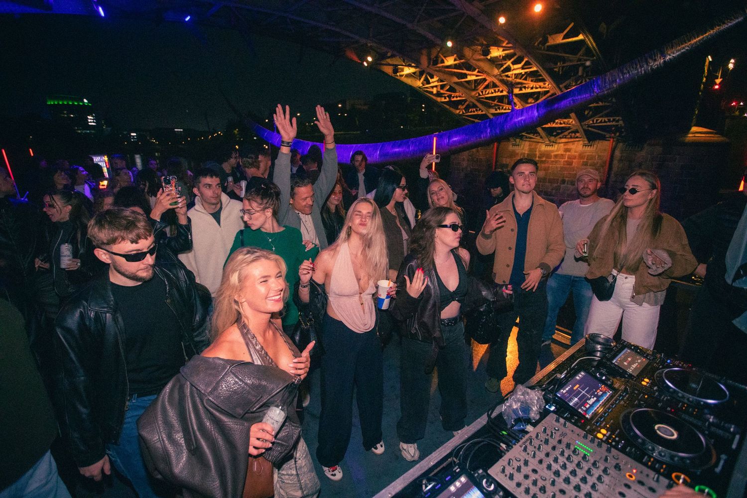 Energetic crowd dancing beside DJ equipment at an indoor nightclub party with purple lighting.