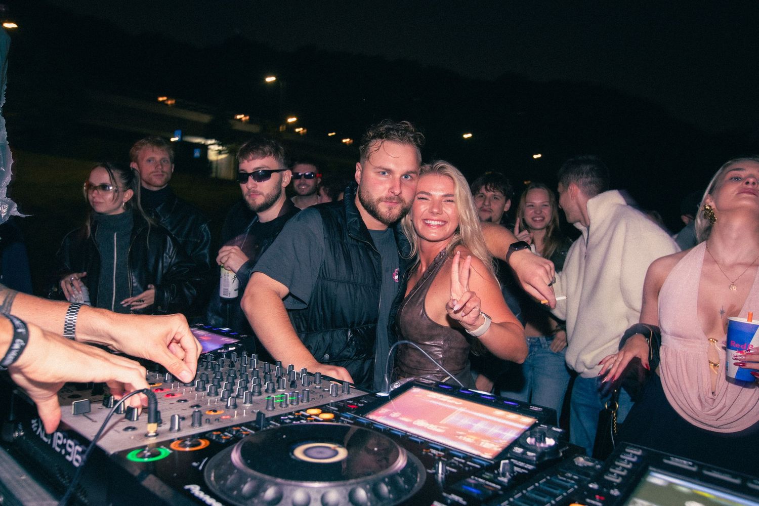 Smiling couple poses together at a DJ booth during a lively outdoor nighttime music event.