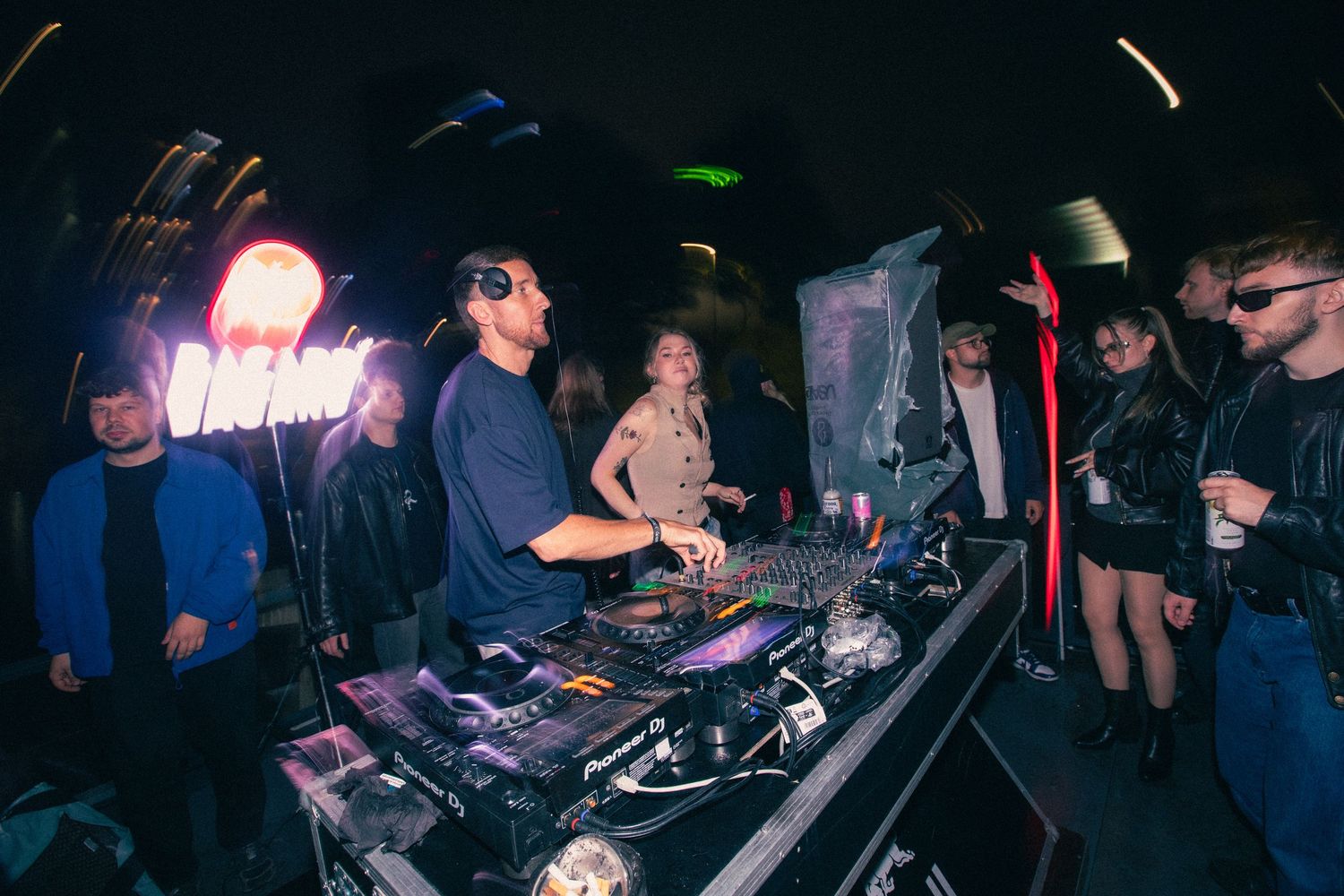 DJ performs at a booth with neon signs while guests watch at a dark indoor nightclub.