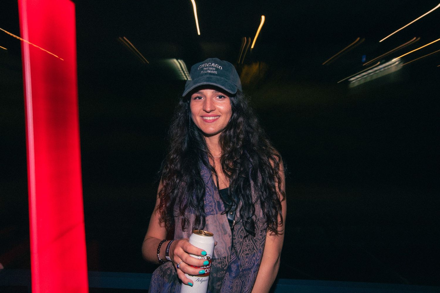 Smiling woman with long curly hair holds a drink near a red light at a nighttime event.