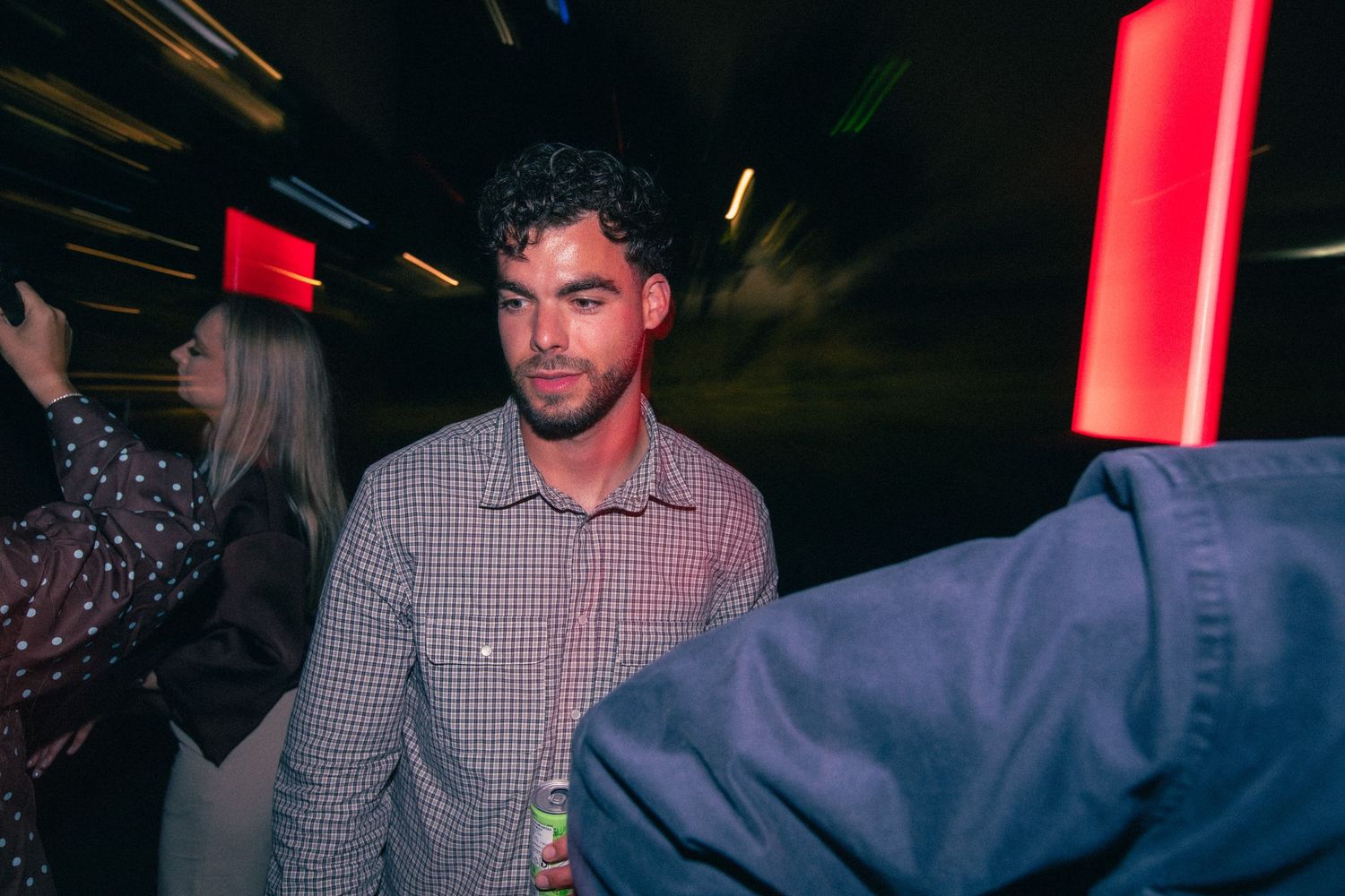 Man in a plaid shirt stands in a dark venue with red light streaks at a night event.