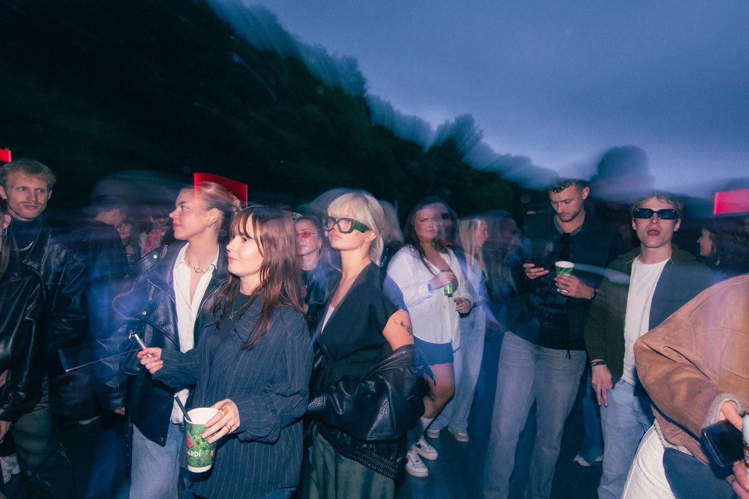 Young crowd gathered at an outdoor evening concert, some wearing glasses and holding drinks.