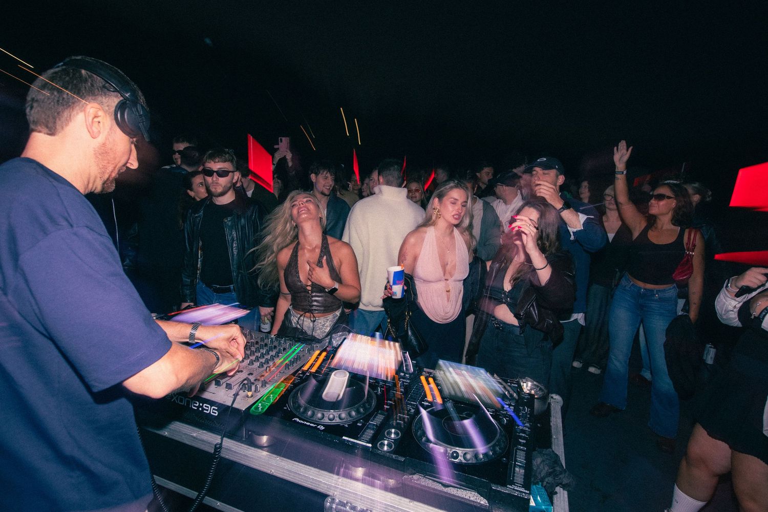 DJ mixing at turntables while an enthusiastic crowd dances at a dark nightclub event.