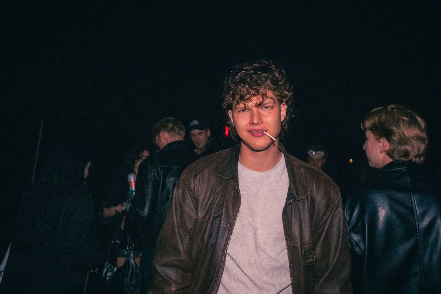Young man in a brown leather jacket stands in a dark crowded nightclub setting.