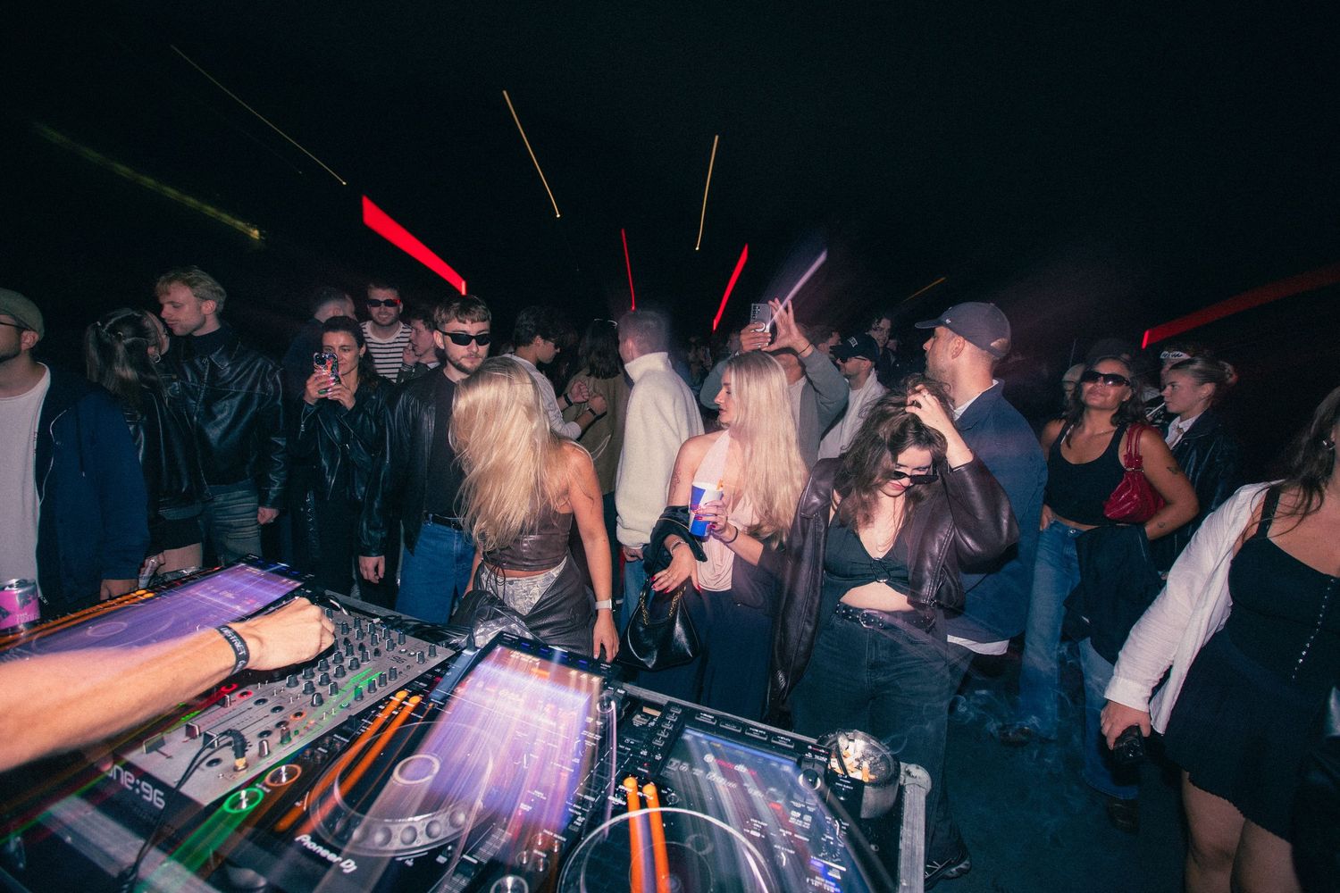 Energetic nightclub crowd dancing near a DJ booth with colorful lights and mixing equipment.