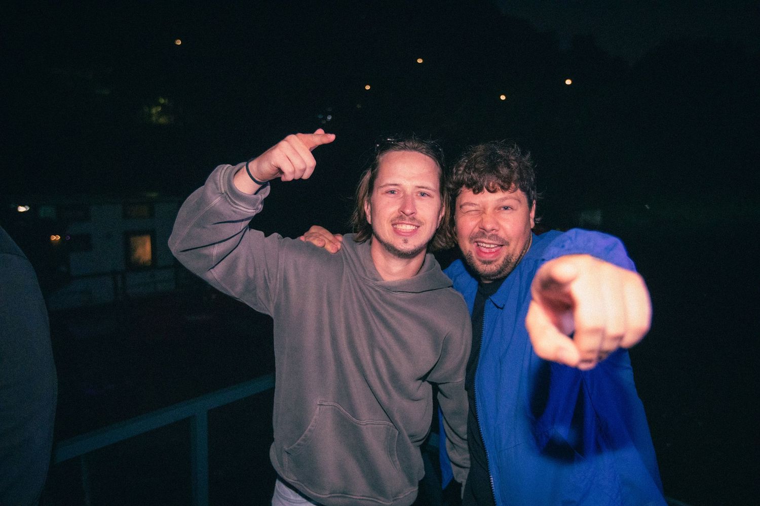 Two smiling men pointing at camera during a nighttime outdoor music festival event.