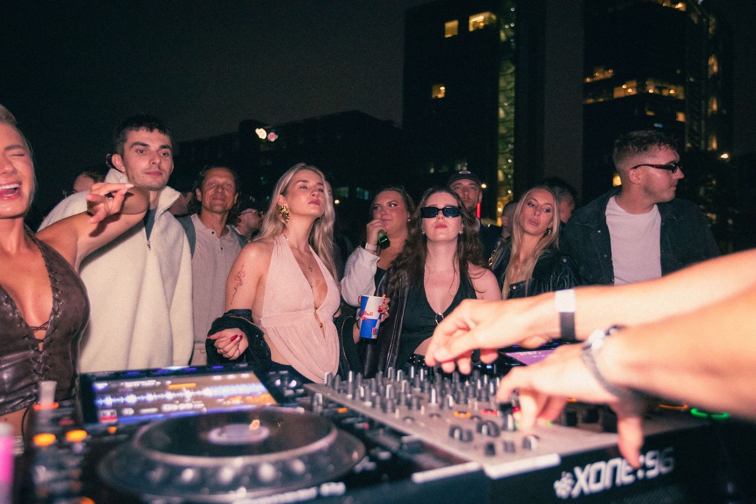 Partygoers dancing near DJ decks at a rooftop nightclub event with city lights behind.