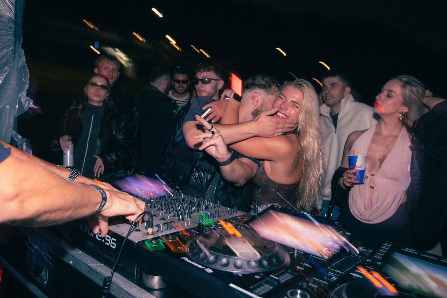 Excited blonde women lean over a DJ booth with colorful mixer at a lively night party.
