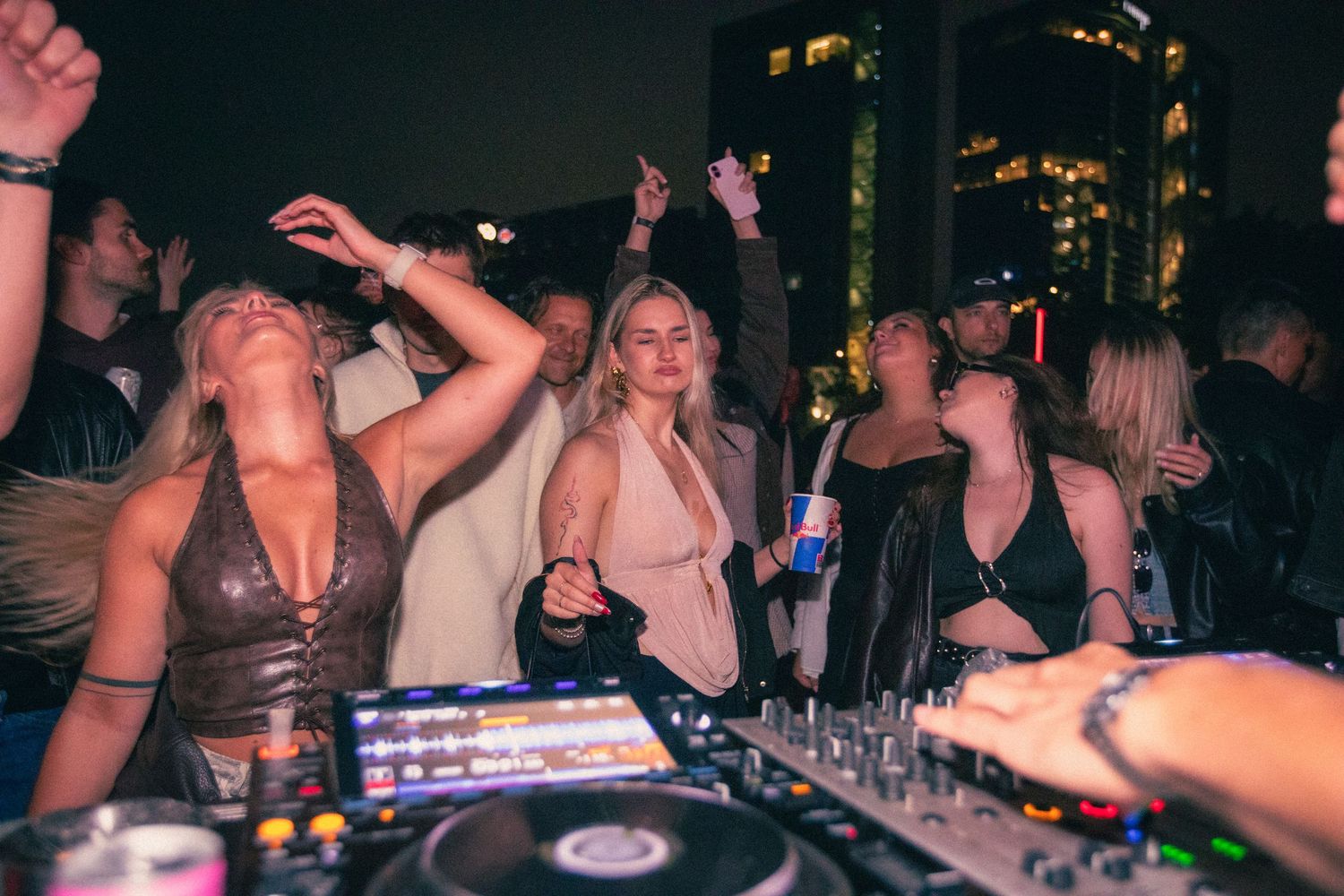 Women dancing near DJ turntables at a lively nightclub party with city skyline backdrop.