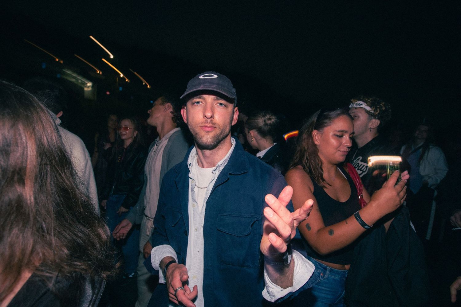 Young man in a blue jacket and cap gestures at the camera during a crowded night event.