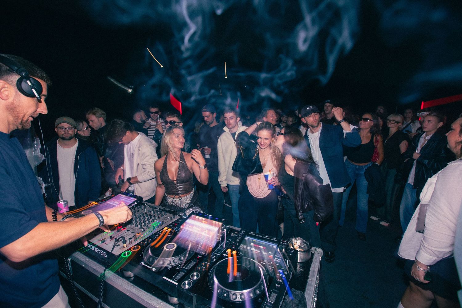DJ mixing at turntables surrounded by dancing crowd at a smoky outdoor nighttime event.