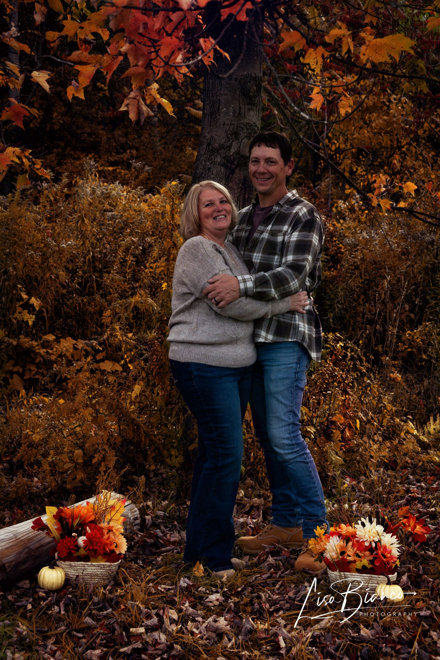 Couple embracing in autumn forest with colorful fall foliage, pumpkins, and floral arrangements during outdoor photo session.
