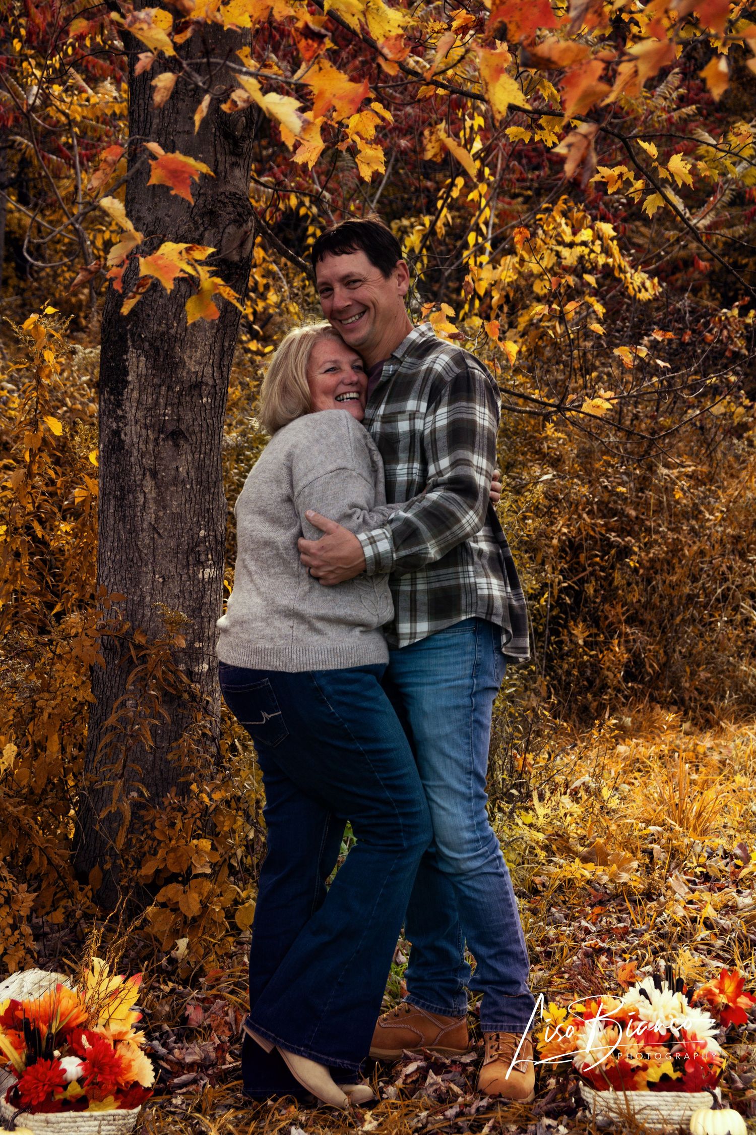 Couple embracing and laughing among vibrant autumn foliage, surrounded by fall flowers and pumpkins in a woodland setting.