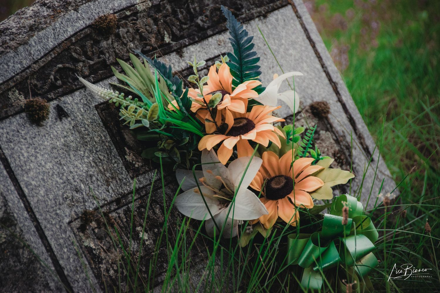 Sunflower and greenery floral arrangement resting on a weathered stone grave marker in a grassy cemetery.