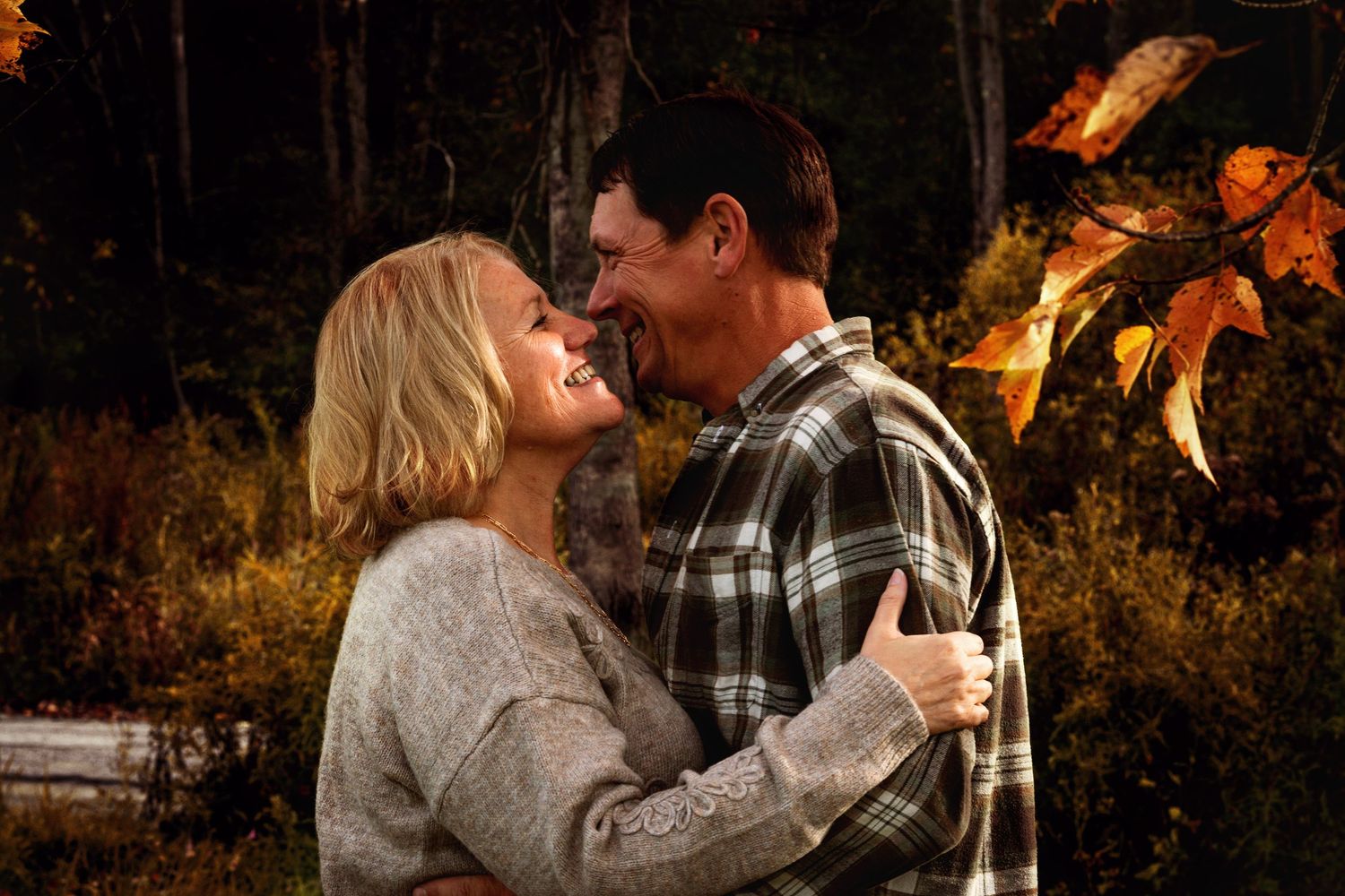 Smiling middle-aged couple embracing in a warm autumn forest, with colorful orange leaves hanging in the foreground.