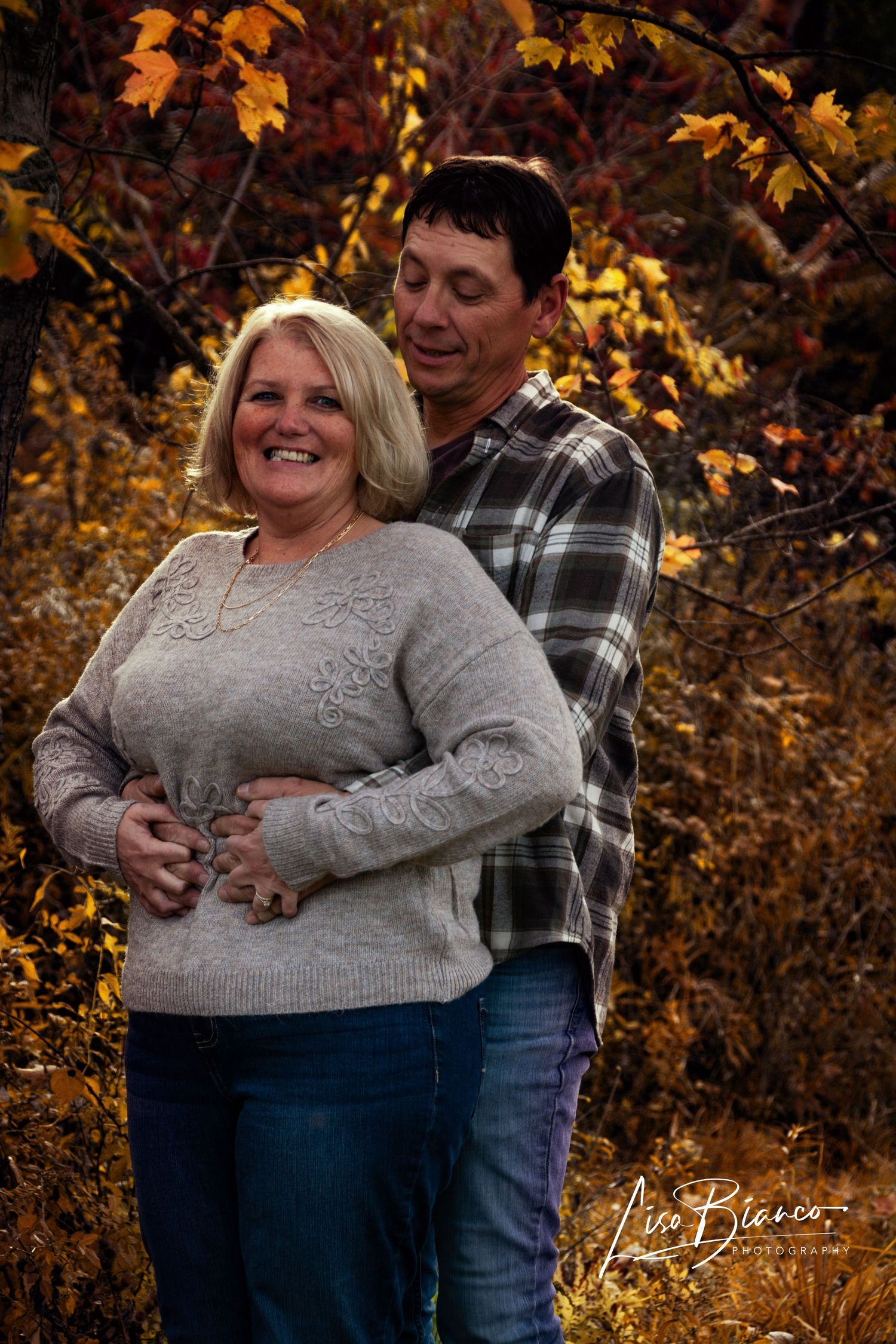 Couple embracing outdoors among vibrant autumn foliage, woman in gray sweater smiling, man in plaid shirt, fall portrait.