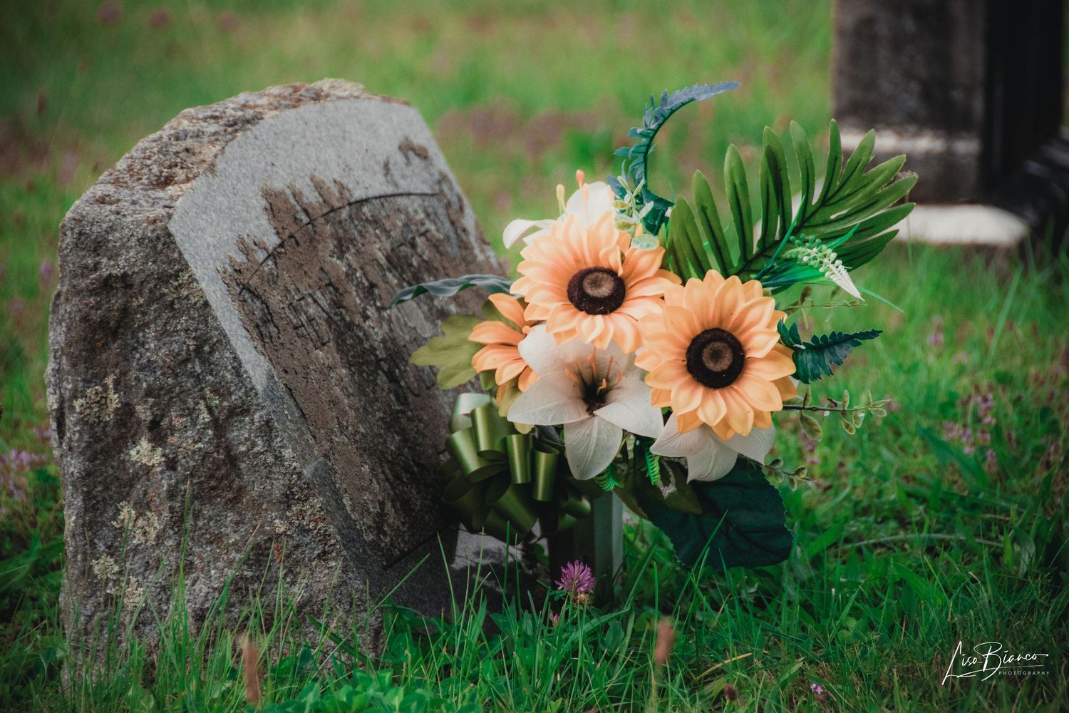Sunflower bouquet with tropical leaves placed beside a weathered stone grave marker on green grass.