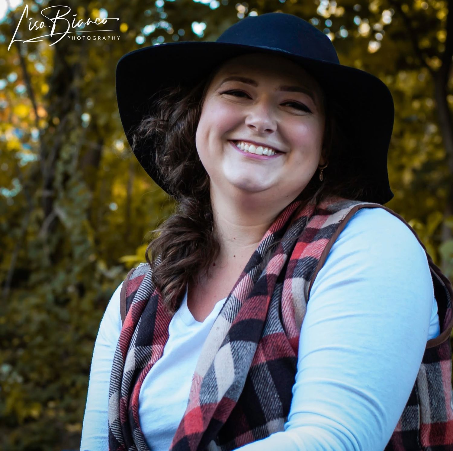 Smiling woman wearing a black wide-brim hat and plaid flannel vest, posing outdoors with autumn foliage background.
