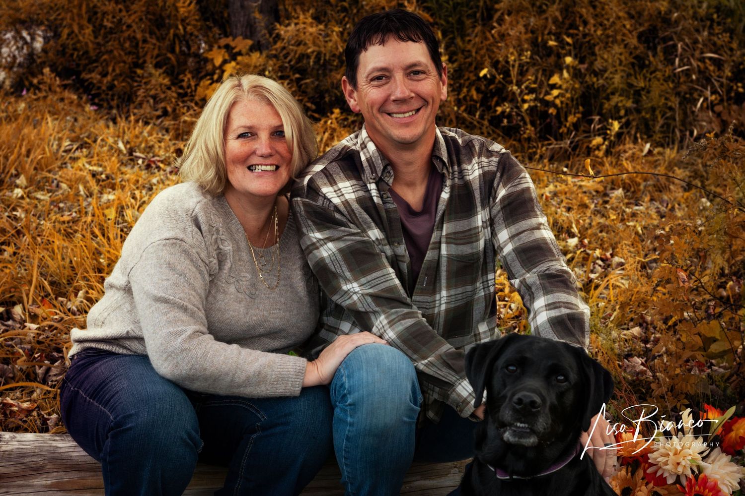 Smiling couple posing outdoors with a black Labrador dog among vibrant autumn foliage in a fall family portrait.