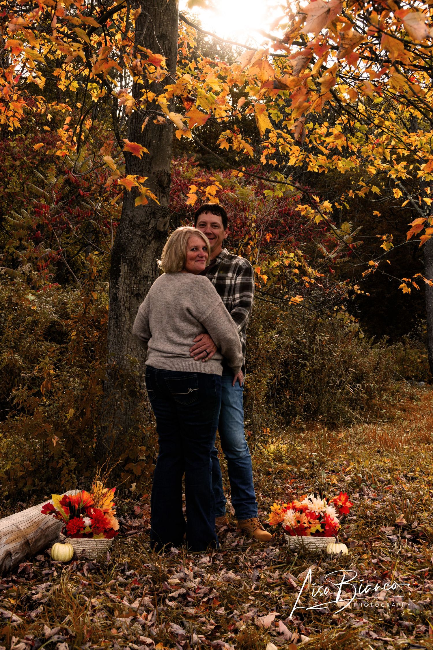 Couple embracing in autumn forest with colorful fall foliage, pumpkins, and flowers during a fall engagement photo session.