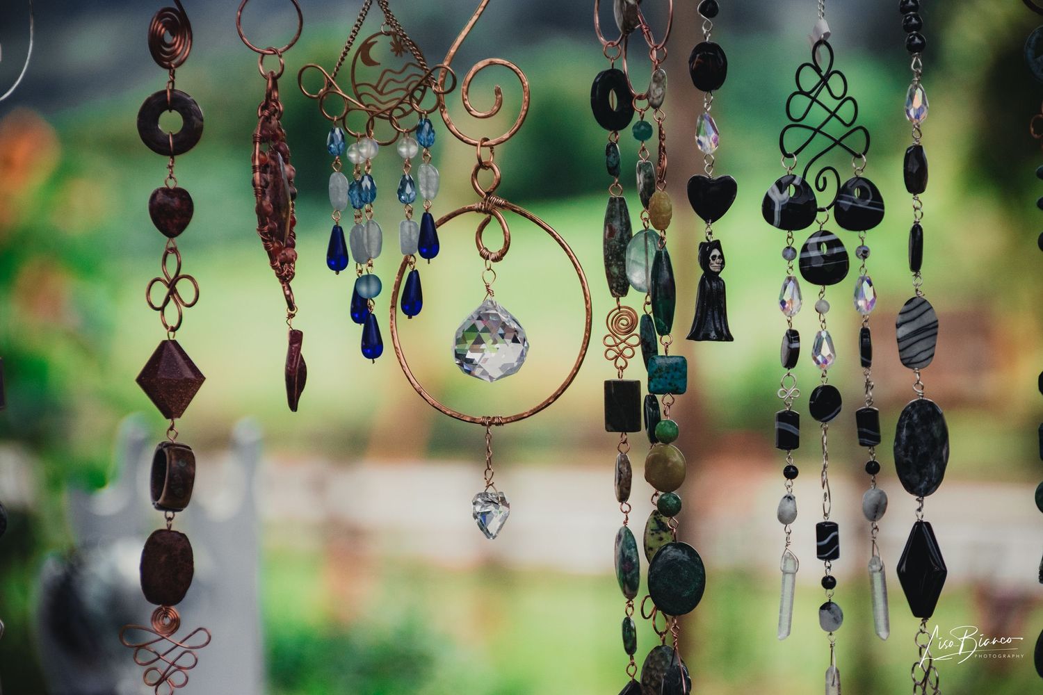 Colorful handmade wind chime sun catchers with crystals, beads, and metal shapes hanging outdoors with blurred green background.