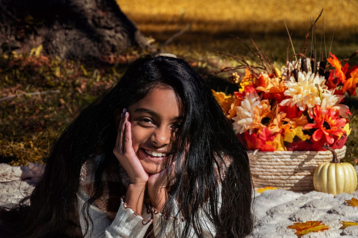 Smiling young woman with long dark hair rests her chin on her hand beside an autumn flower basket and small white pumpkin outdoors.