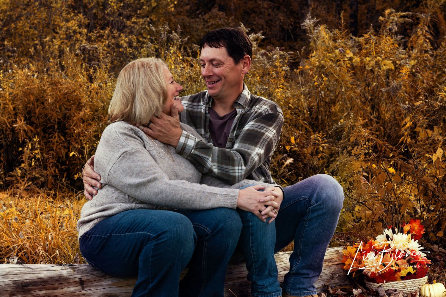 Couple laughing together outdoors in fall foliage, sitting on a log in a warm autumn setting.