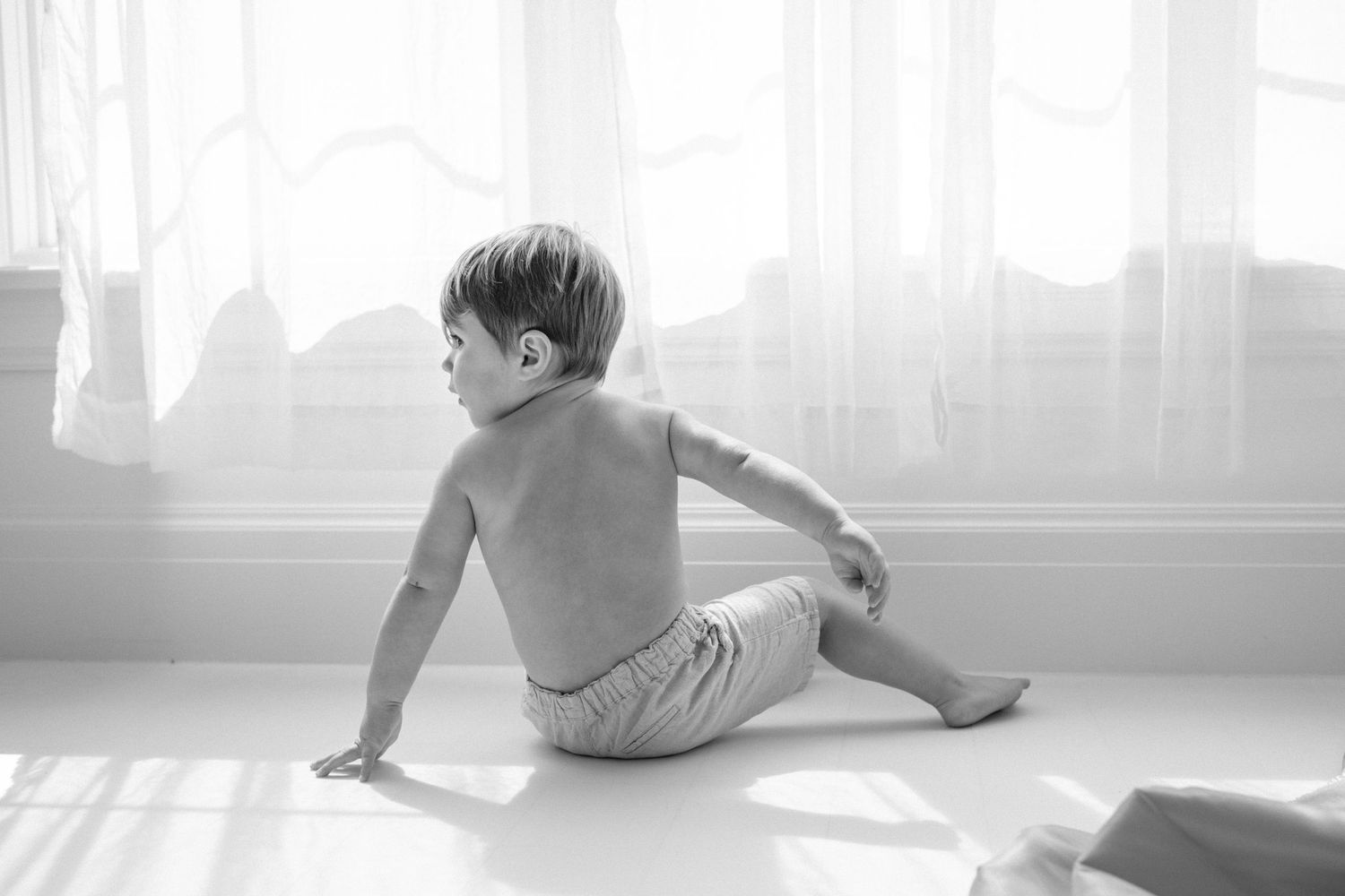 Black and white photo of a shirtless toddler sitting on a white floor, looking toward a bright sunlit window.