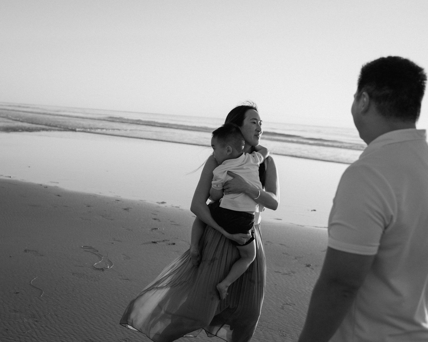 Black and white beach photo of woman in flowing dress carrying child while man watches at sunset.