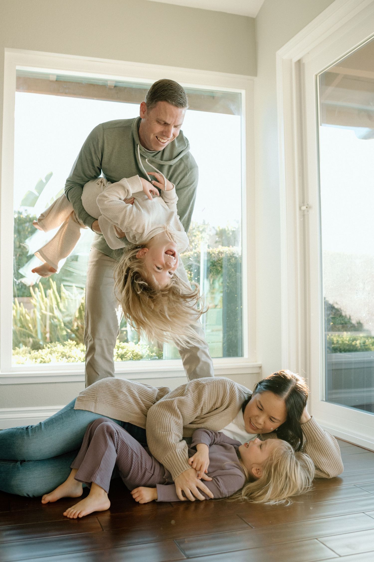 Playful family of four with dad holding child upside down while mom and kids laugh on floor.