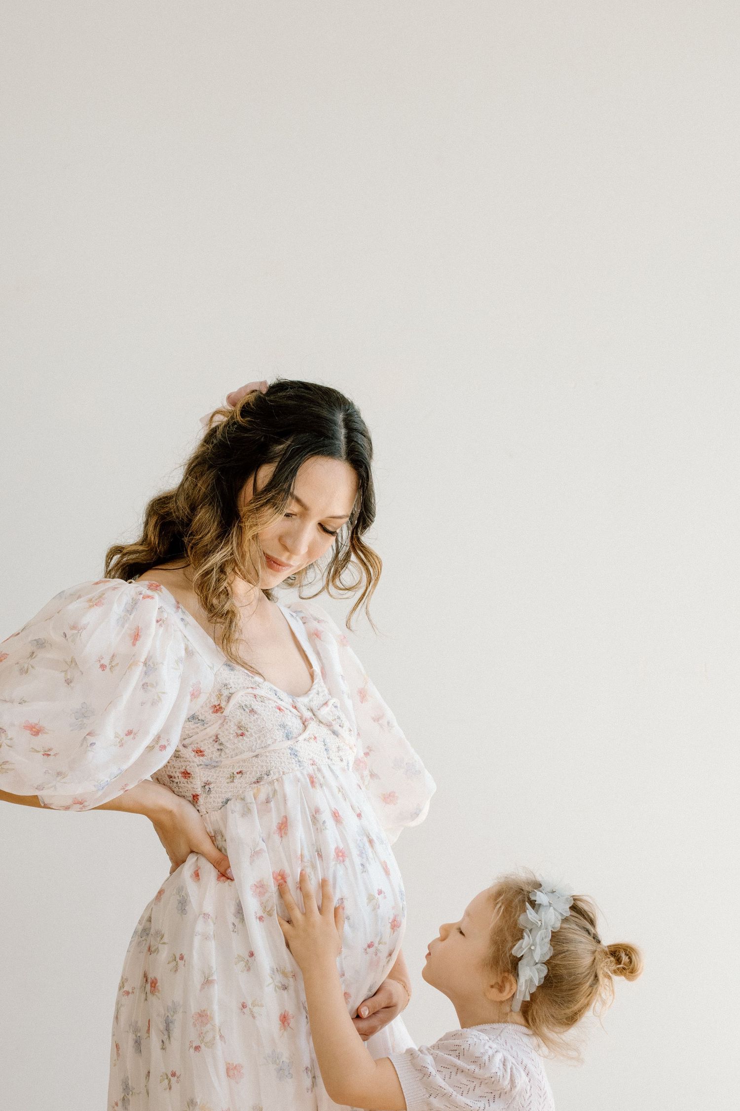 A pregnant woman in a floral dress looks down as her young daughter gently touches her baby bump.
