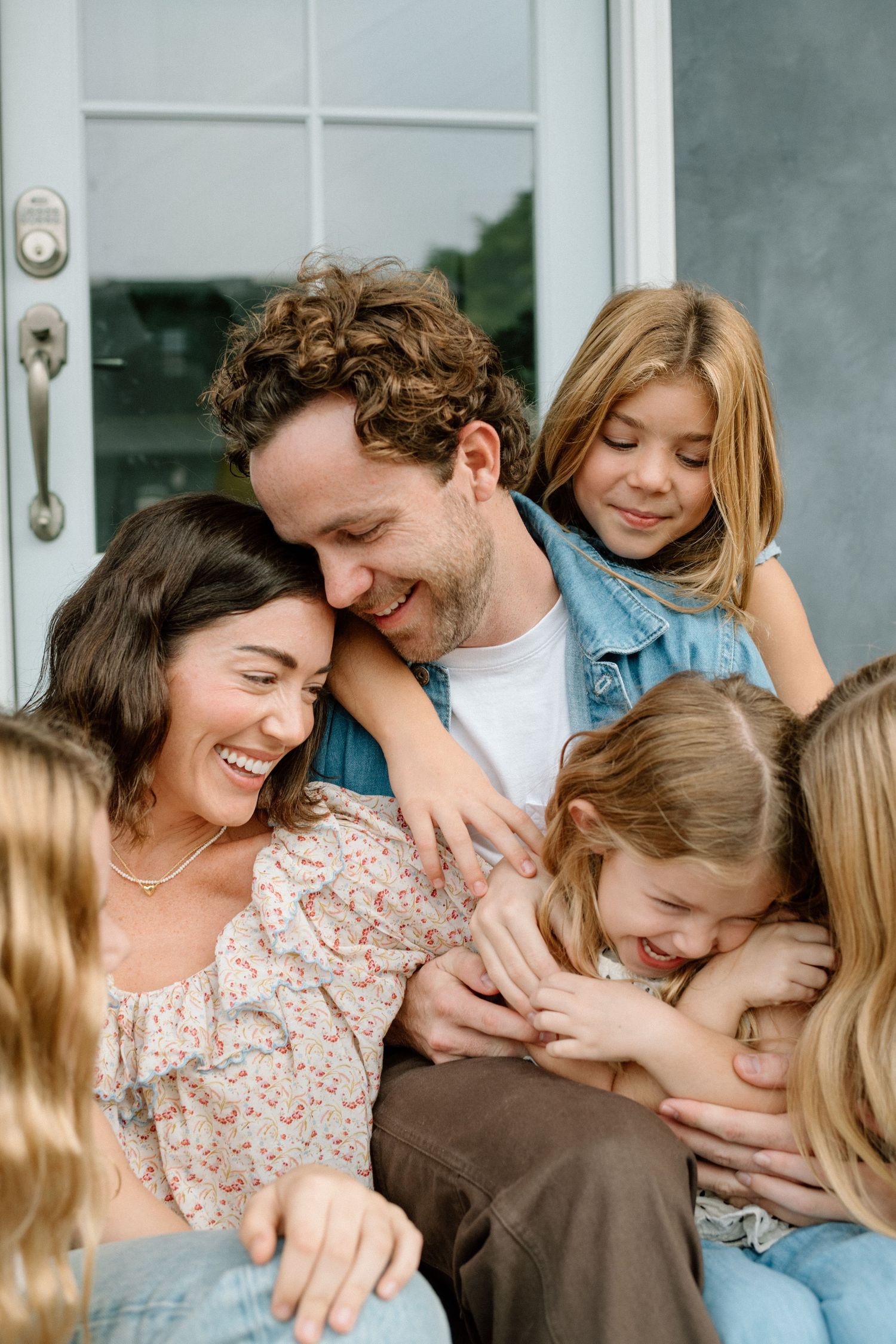 Happy family laughing and cuddling together on front porch steps in casual clothing.