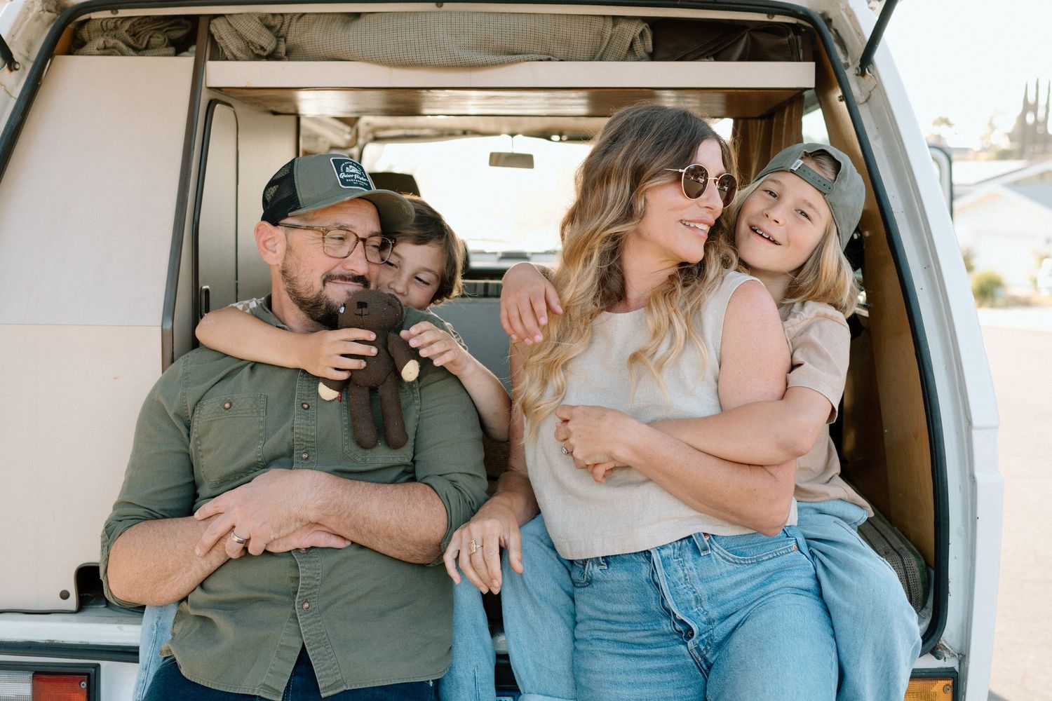 Happy family of four sits in open camper van trunk, smiling and embracing outdoors.
