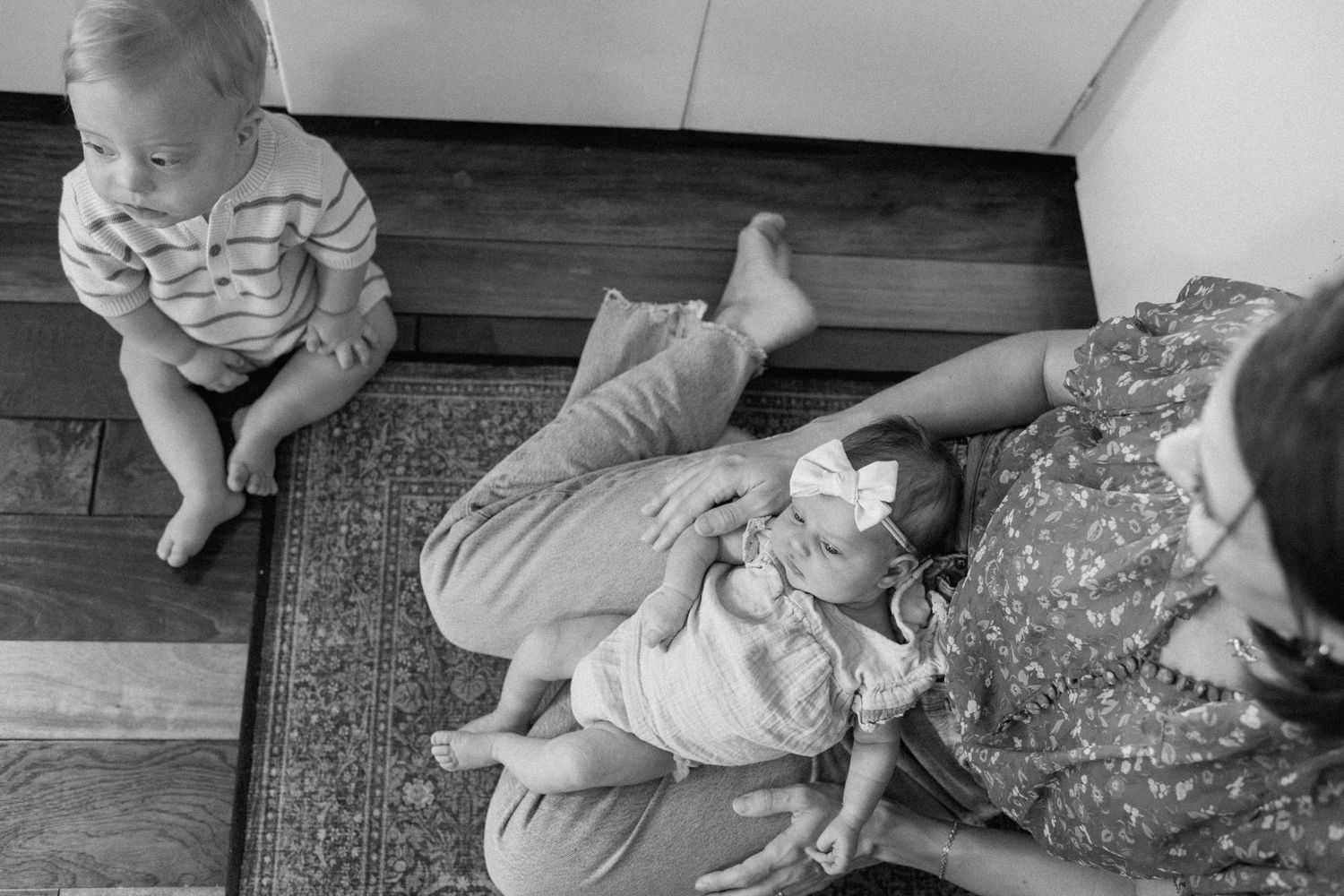 Black and white photo of a mother cradling her newborn baby girl while a toddler sits nearby on a hardwood floor.