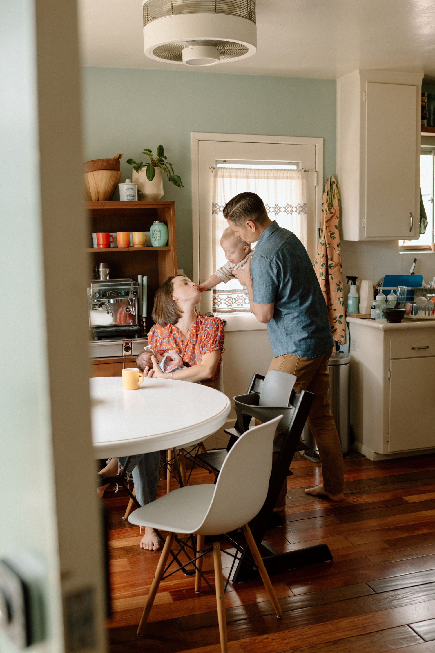 A man stands holding a baby while a woman sits at a white round kitchen table in a cozy home.