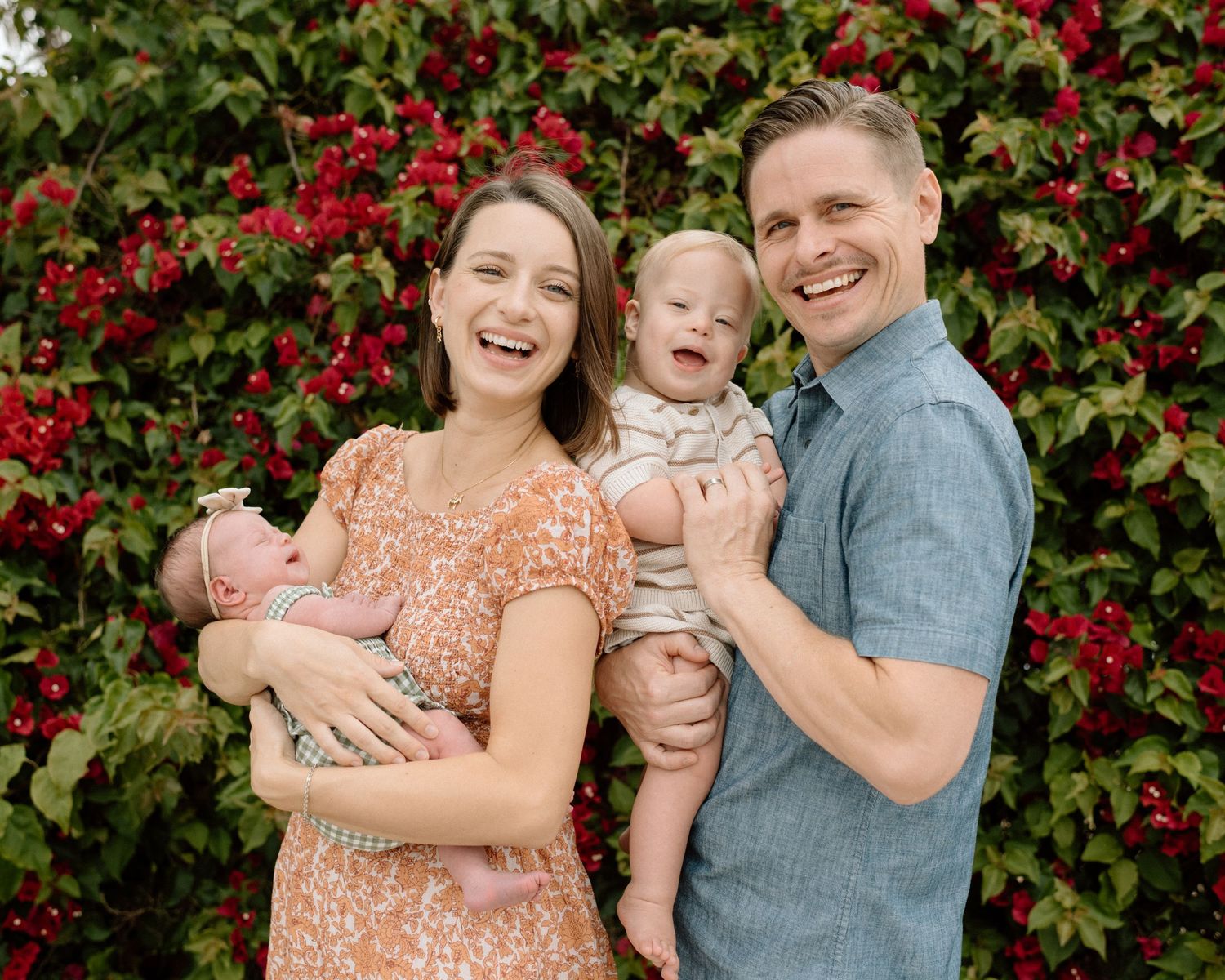 Happy family of four posing outdoors with two babies in front of vibrant red flowering bushes.
