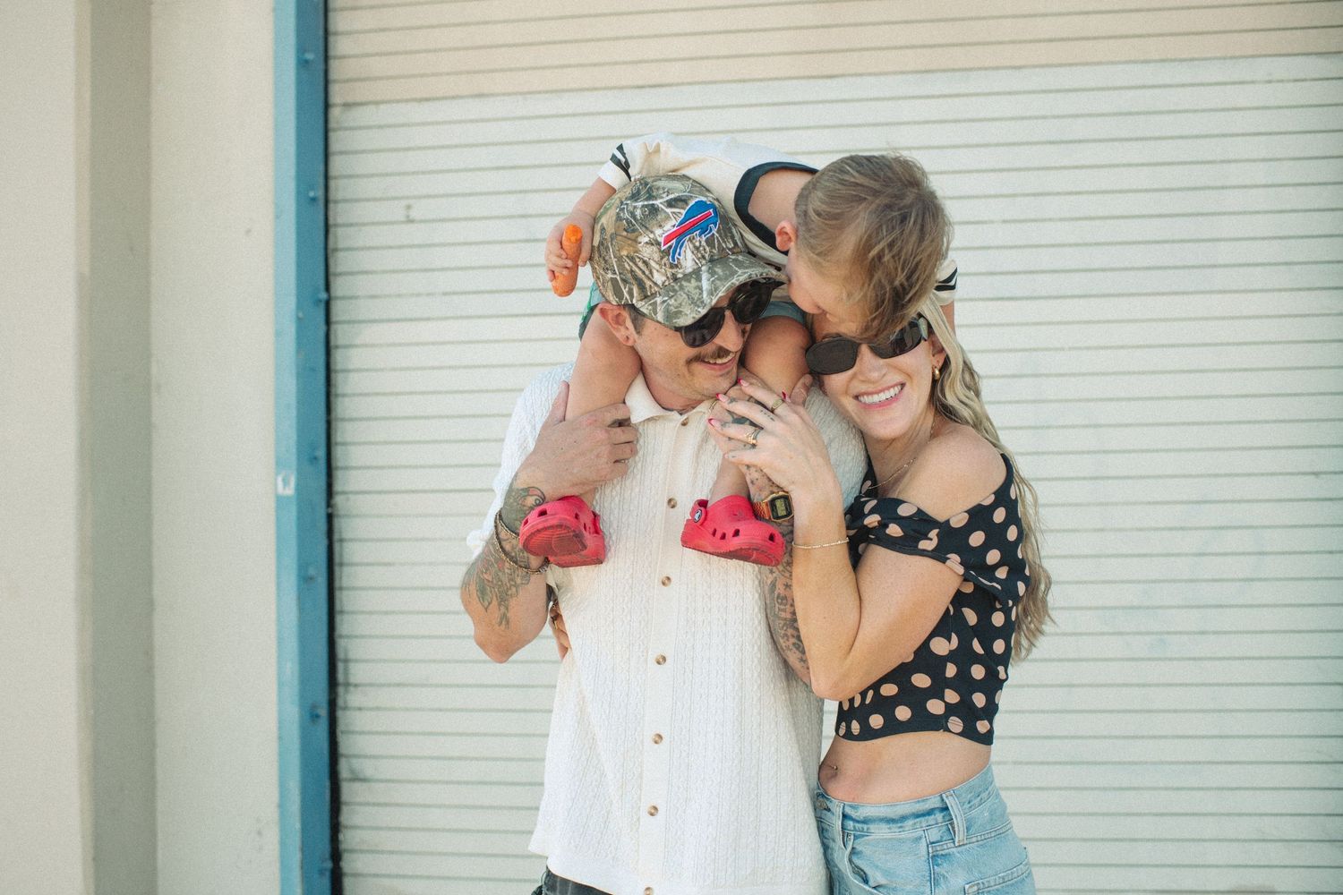 Tattooed man in colorful cap and sunglasses laughing with two women outdoors near a white garage door.