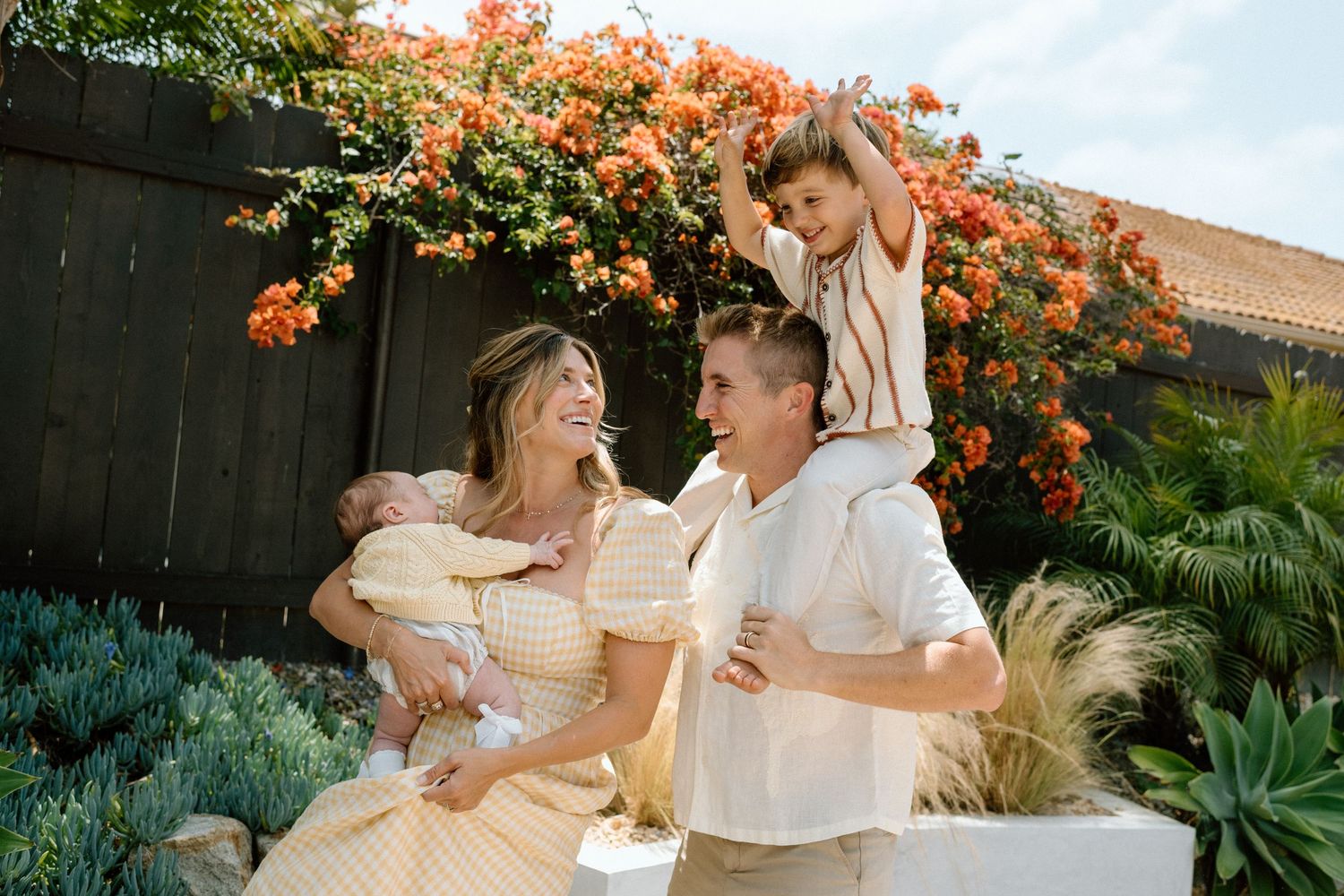 Happy family with two young children posing in a sunny garden with vibrant orange bougainvillea flowers.