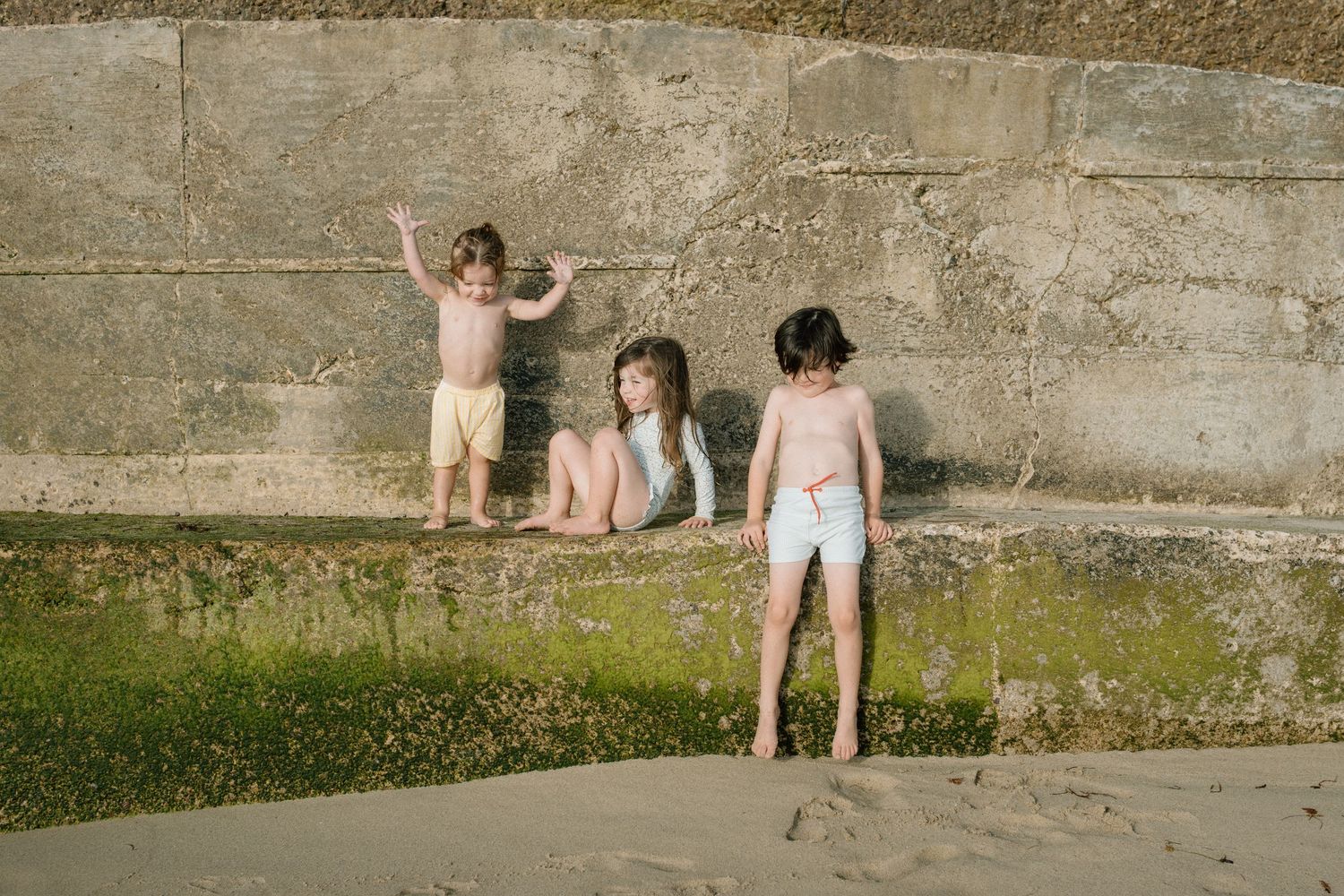Three young children playing near a stone wall on a sandy beach in summer.