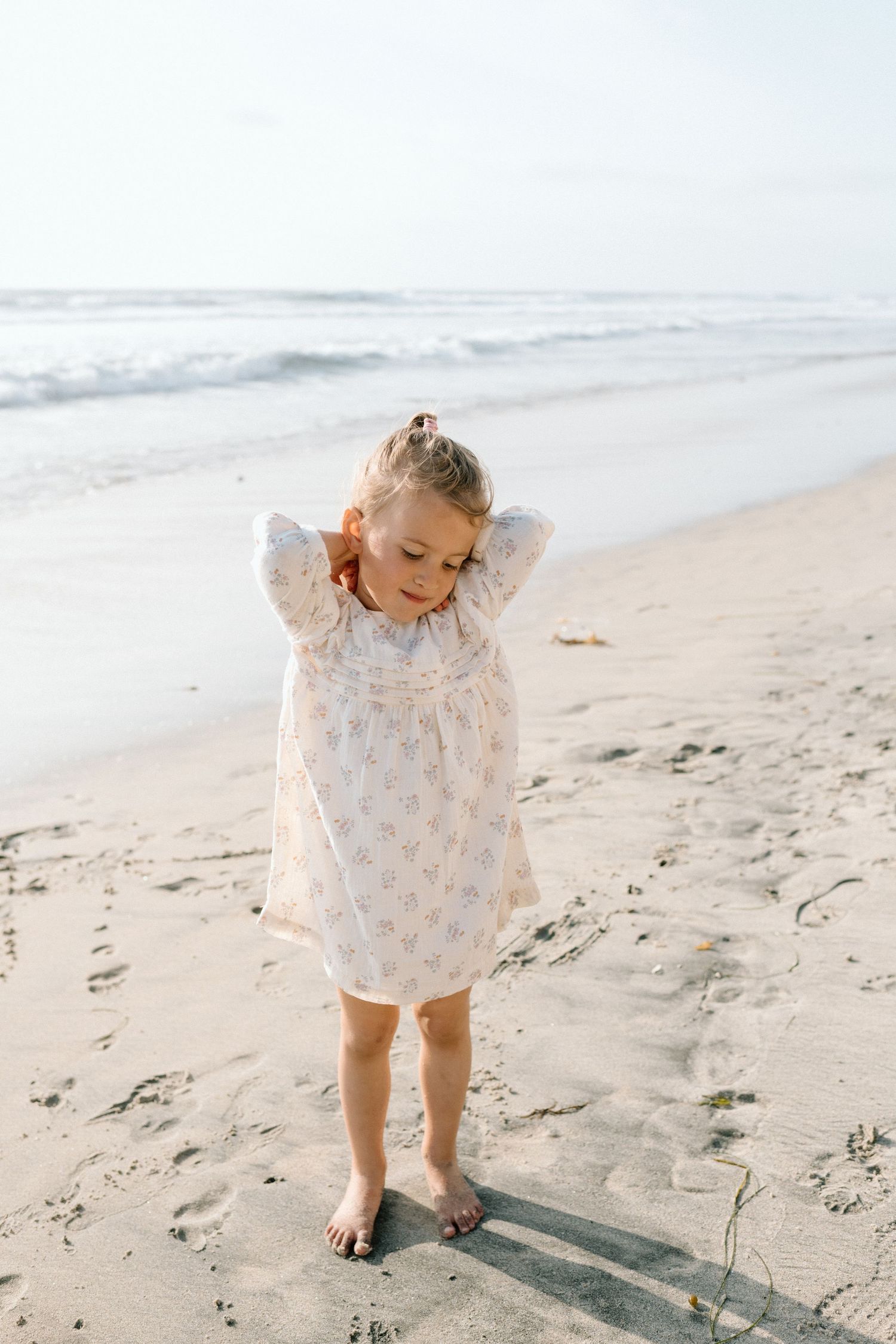 Young girl in floral dress standing barefoot on sandy beach with ocean waves in background.