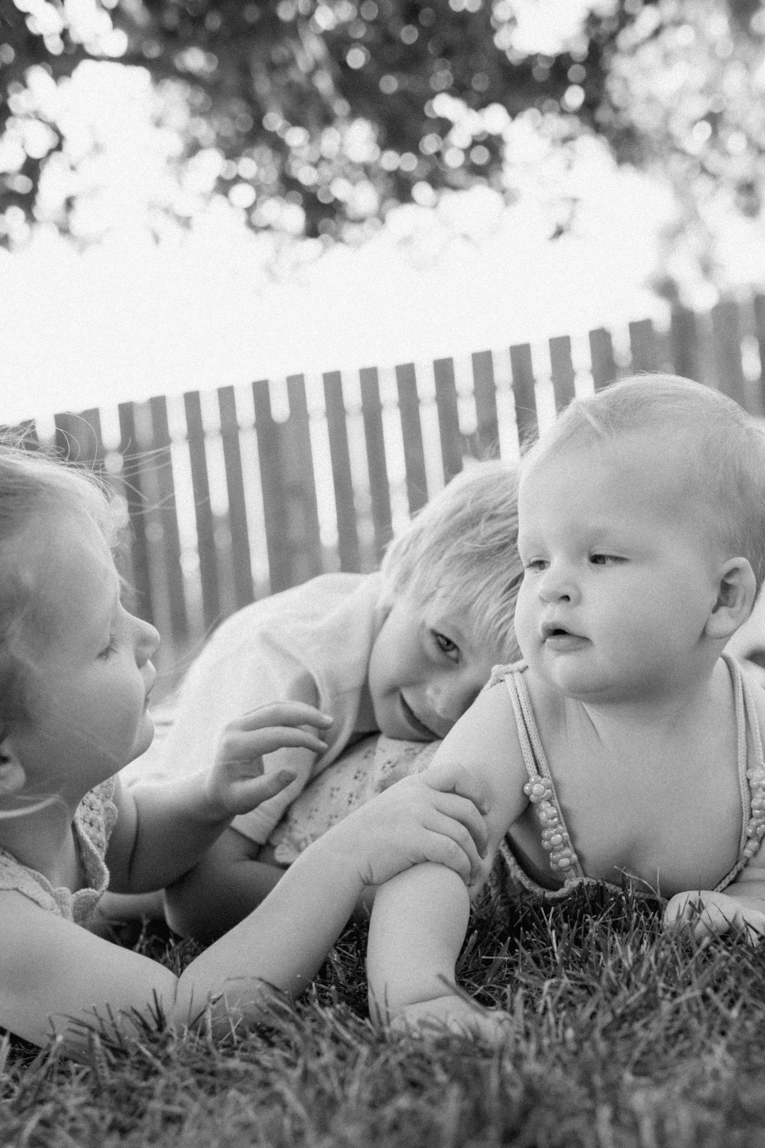 Black and white photo of three young siblings playing together on grass in backyard.