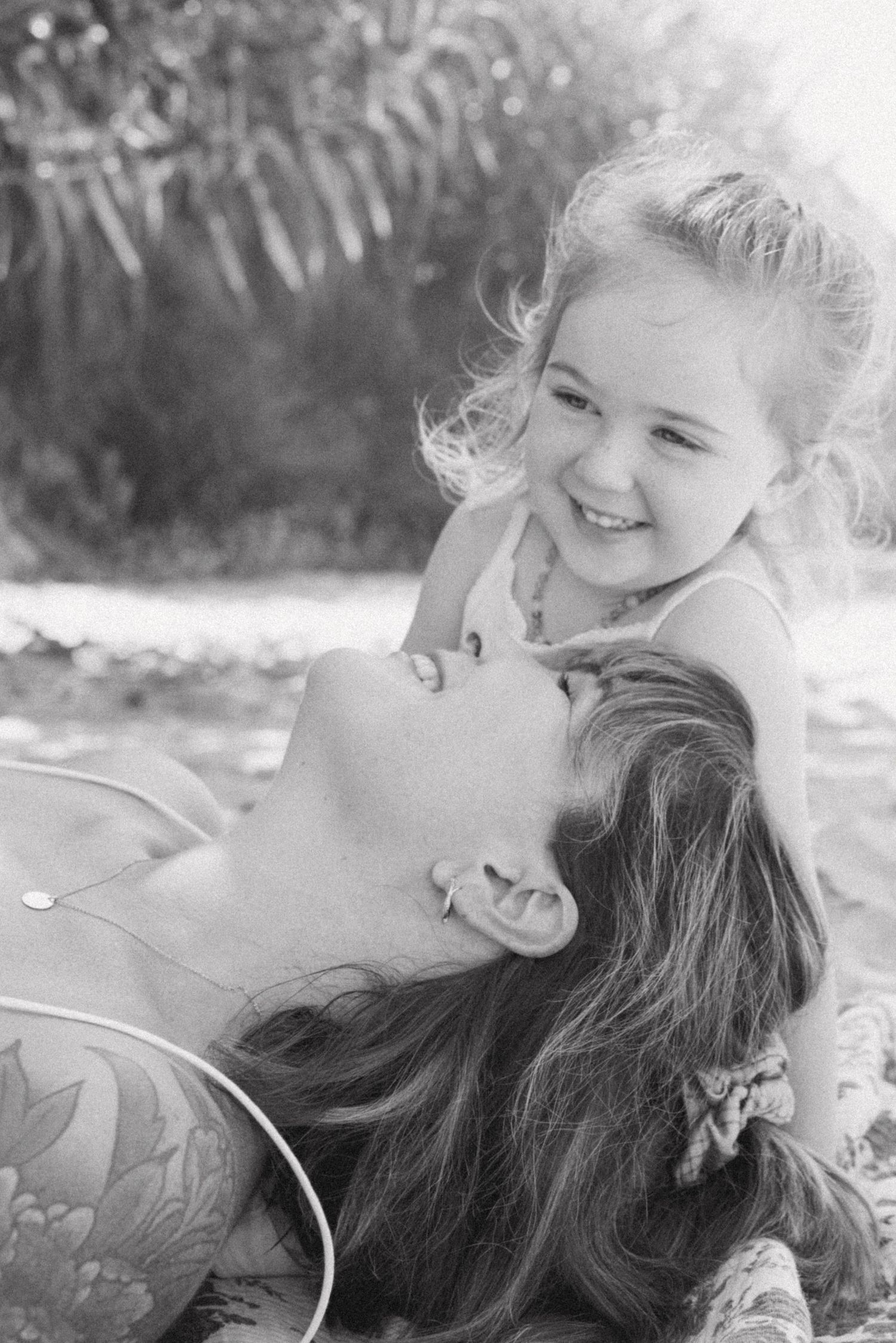 Black and white photo of a laughing child leaning over a smiling woman lying down outdoors.